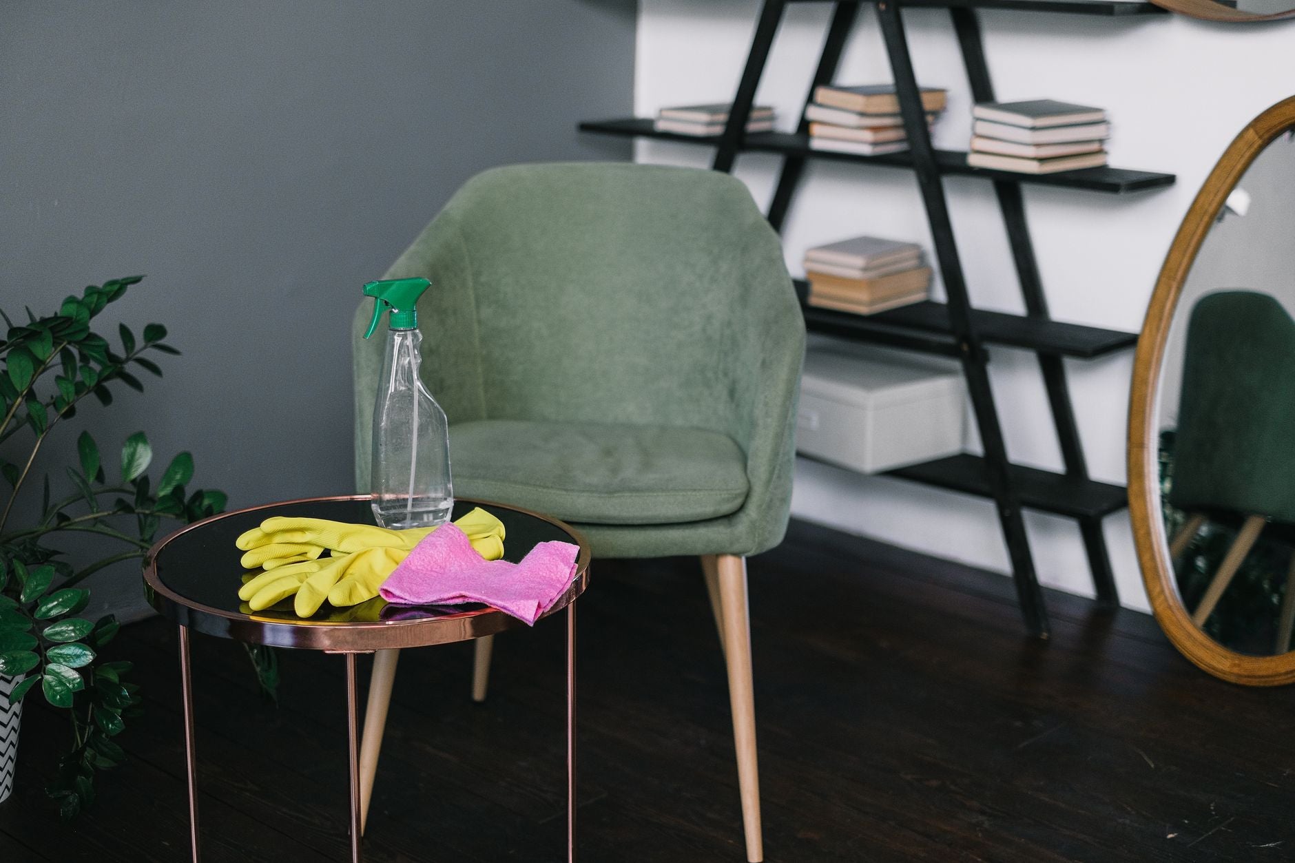 Yellow latex gloves placed near spray detergent bottle and rag on round table against comfortable chair in living room in...